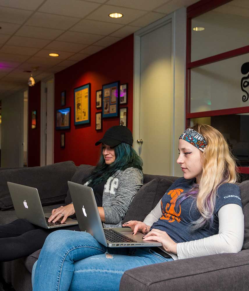 Two female emplyees at Turbosquid sit comfortably on couches while working on their laptops
